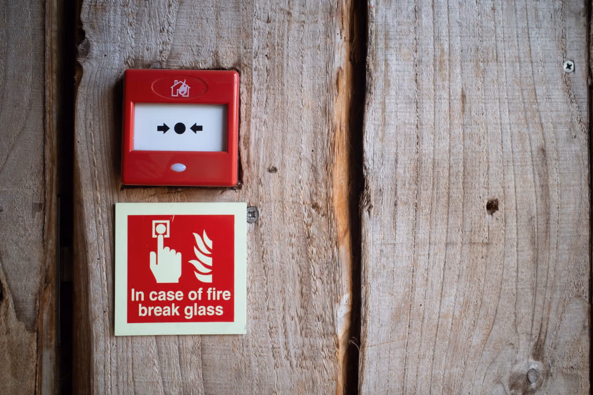 A red manual fire alarm call point mounted on a timber wall above an "In case of fire break glass" sign.