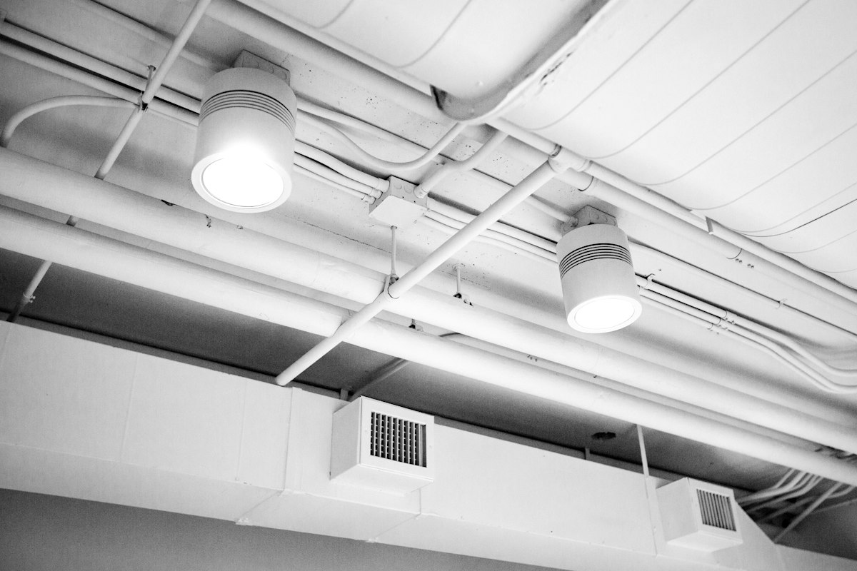 Black-and-white view of an exposed ceiling with white ductwork, supply grilles, pipework and two cylindrical light fittings.