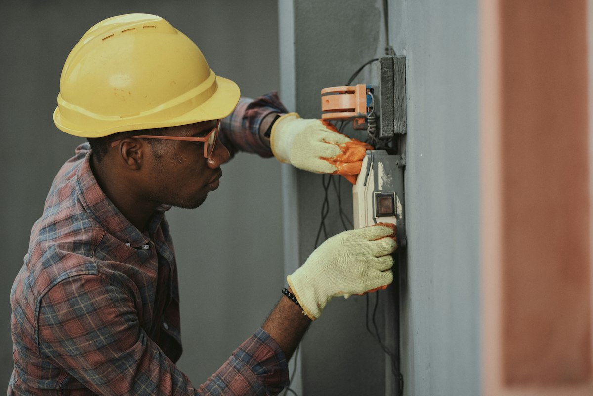 An electrician wearing a yellow hard hat, safety glasses and work gloves fits wiring into an electrical box mounted on a wall.