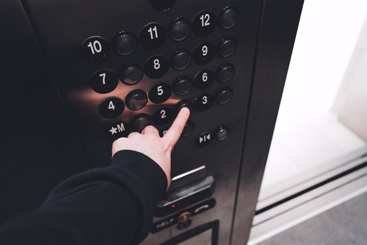 A finger pressing a button on a lift car's floor selection panel, with a HELP button and handset symbol at the bottom of the panel.