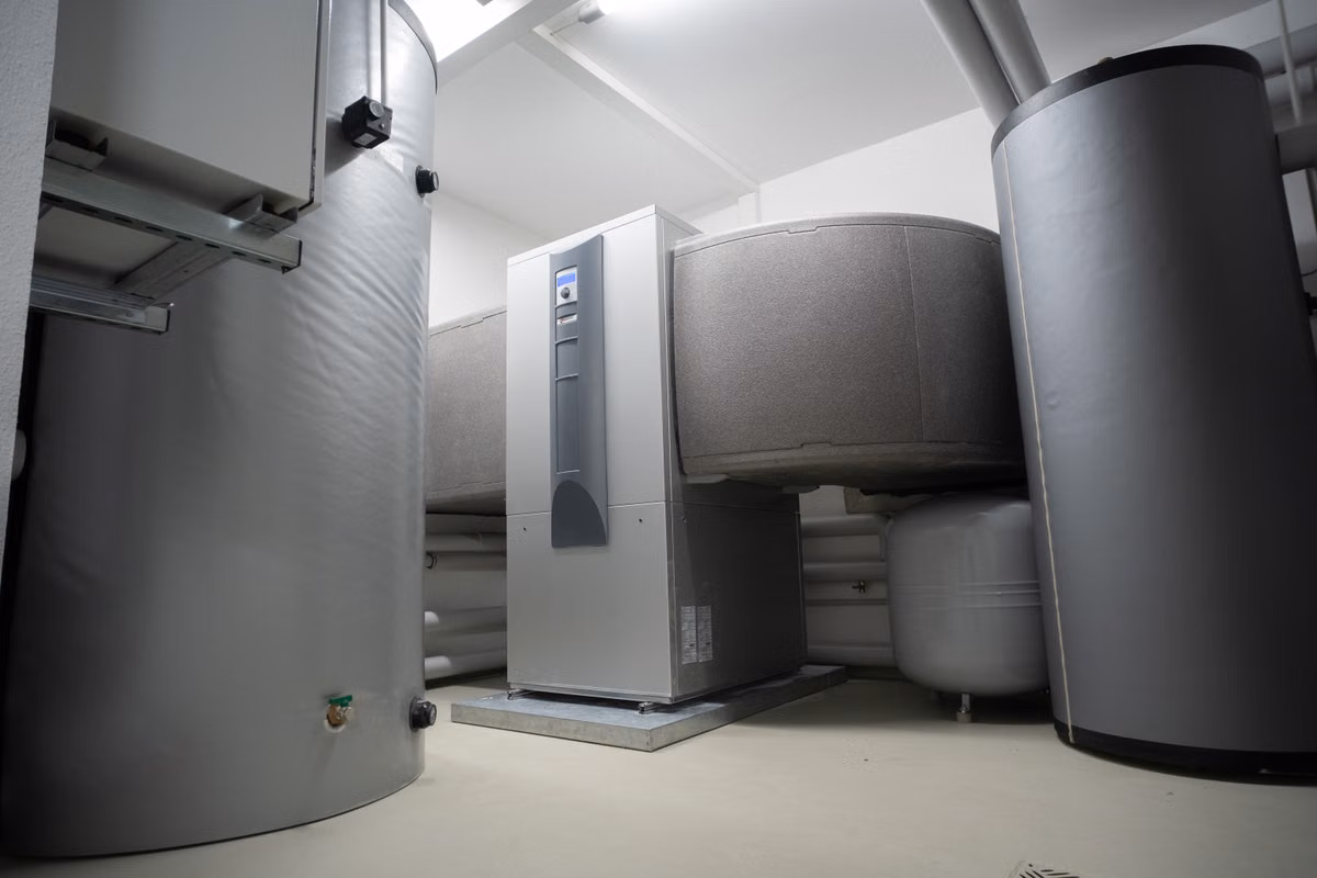 A plant room containing a floor-standing heat pump unit flanked by large insulated hot water storage cylinders, with pipework and an expansion vessel behind.